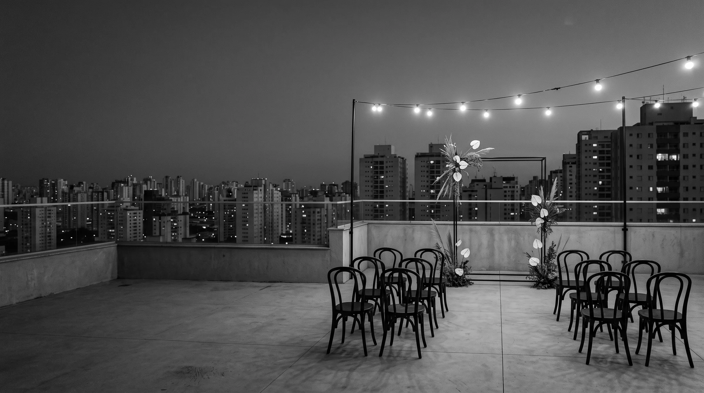 Rooftop de concreto com cadeiras pretas, arco de aço e o skyline de São Paulo à noite, em preto e branco